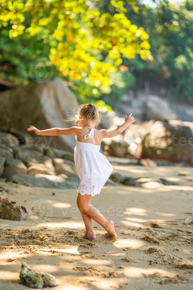 pequeño niña en blanco vestir teniendo divertido en costa en el sombra de arboles y palmas alto calidad foto