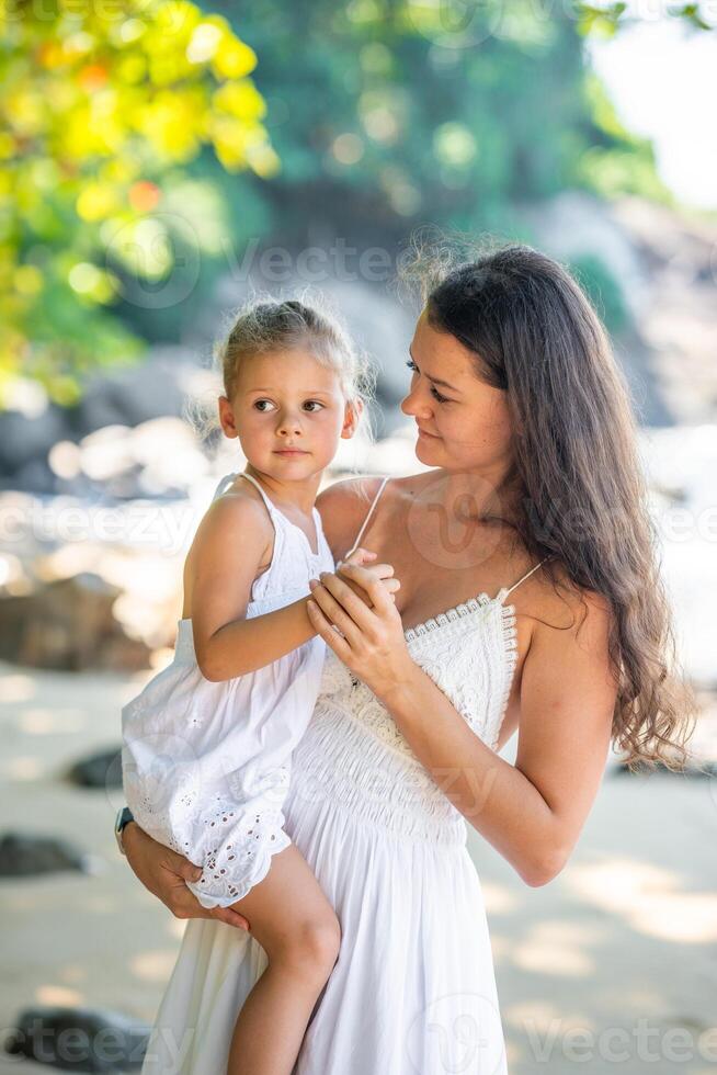 joven mujer madre con un pequeño hija en blanco vestidos en costa en el sombra de arboles y palmas alto calidad foto