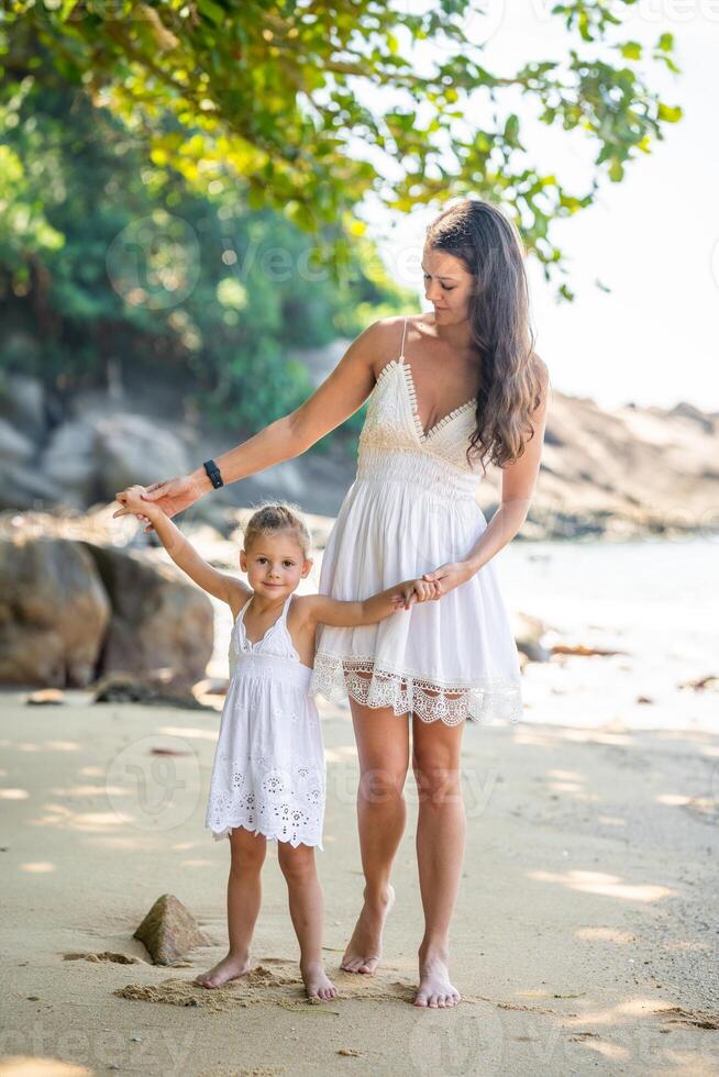 joven mujer madre con un pequeño hija en blanco vestidos teniendo un divertido en costa en el sombra de arboles y palmas alto calidad foto