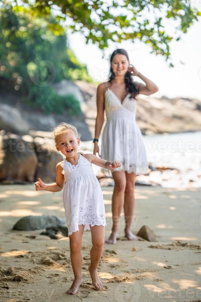 joven mujer madre con un pequeño hija en blanco vestidos teniendo un divertido en costa en el sombra de arboles y palmas alto calidad foto