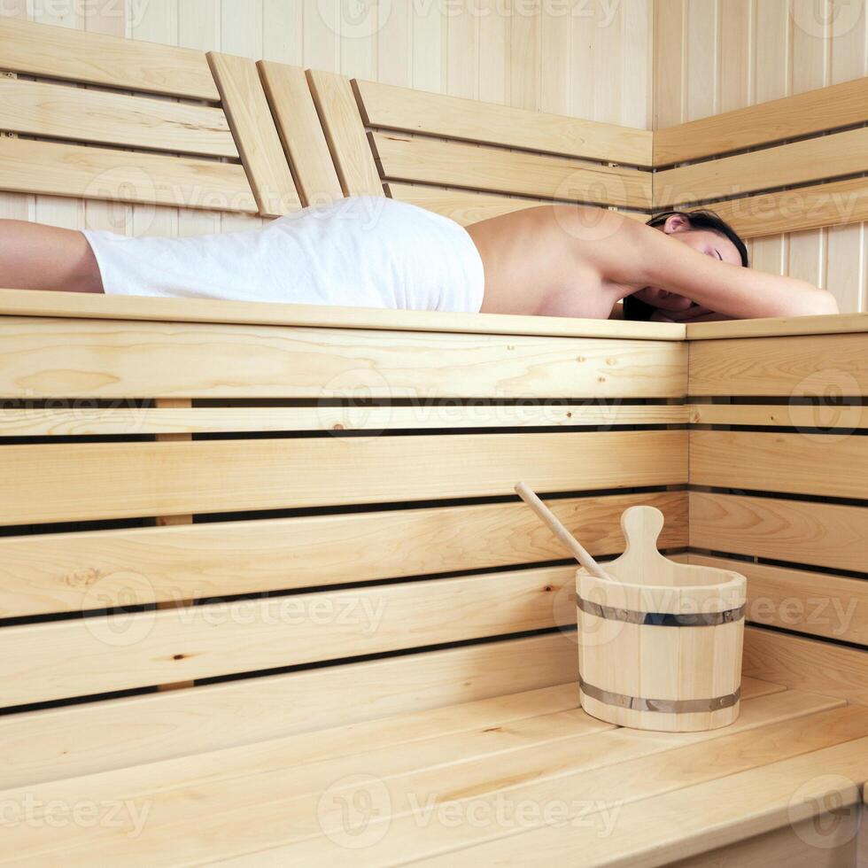 Slim woman wrapped in white towel lies on the bench in a traditional sauna photo