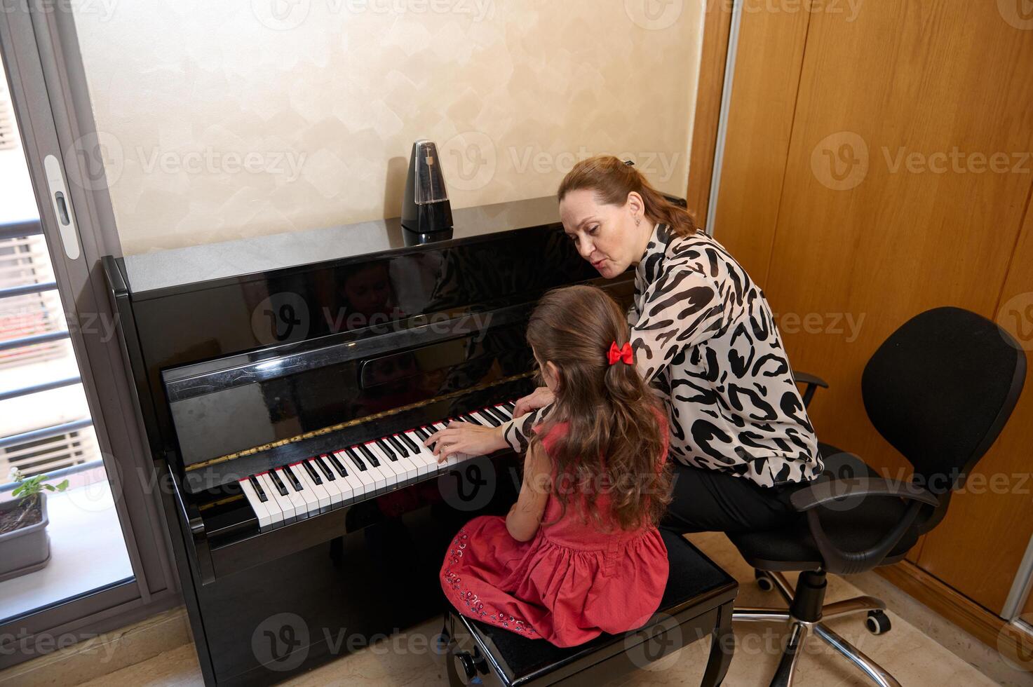 Rear view of a cute little girl in elegant red dress, sitting at wooden piano, with teacher learning to play piano indoors. Musical education and talent development in progress photo