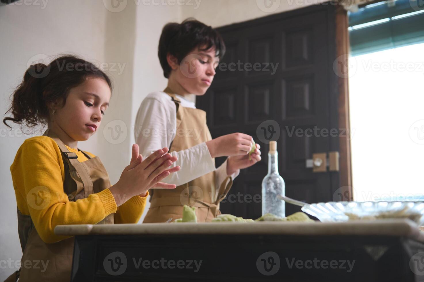 Kids modeling dumplings, standing at floured table at home kitchen interior. Adorable brother and sister preparing a family dinner together, sculpting varennyki according to traditional recipe photo