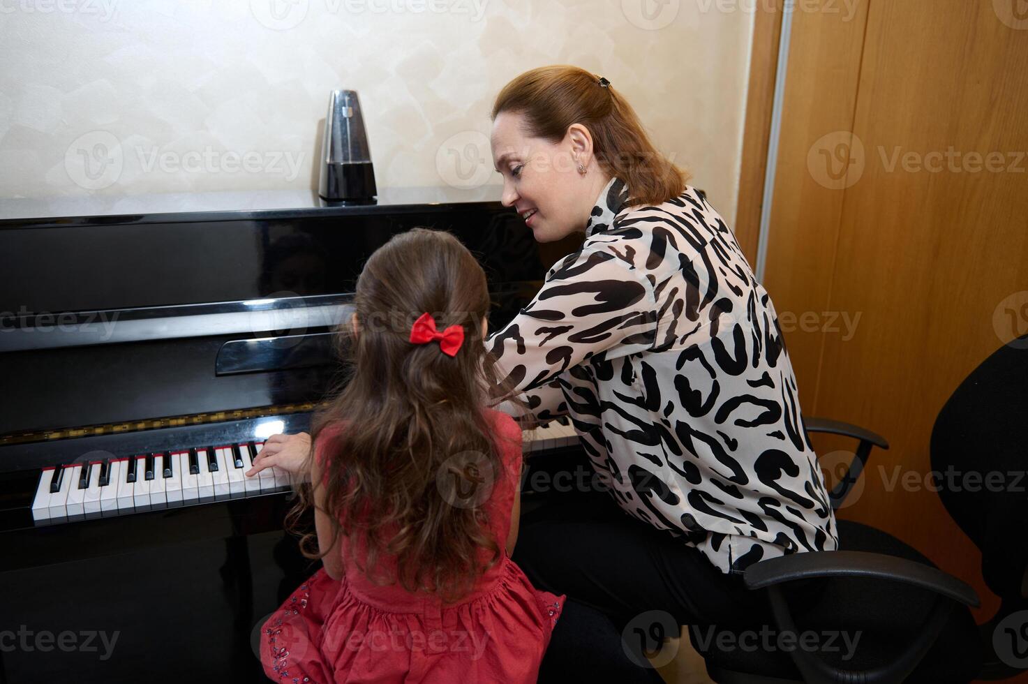 Inspired happy woman pianist, music teacher performing melody on piano forte, explaining piano lesson to a child girl, sitting nearby on a stool. Musical education and talent development in progress photo