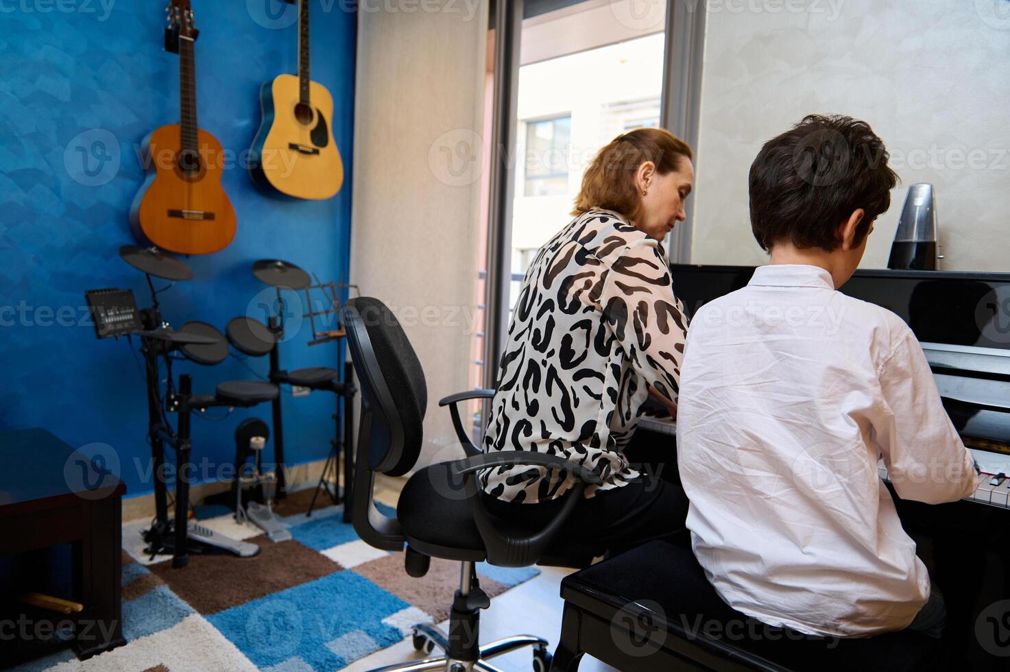 Rear view of a teenage student boy playing piano, composing a melody under his teacher's guidance during individual music lesson at home. Children. Education. Lifestyles. Hobbies and entertainment. photo