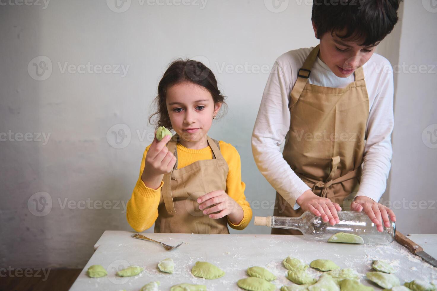 Cute little girl stuffing and molding dough while her brother using wine bottle as rolling pin, rolling out a raw green dough. Kids making ravioli, varennyki, pelmeni against white wall background photo