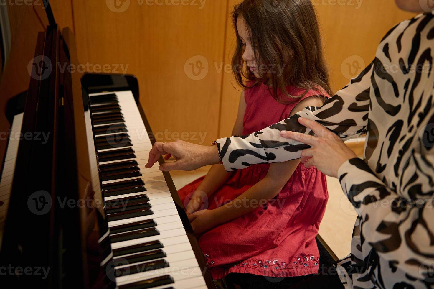 Little kid girl in elegant red dress, taking piano lesson, passionately playing the keys under her teacher's guidance, feeling the rhythm of music. Musical education and talent development in progress photo