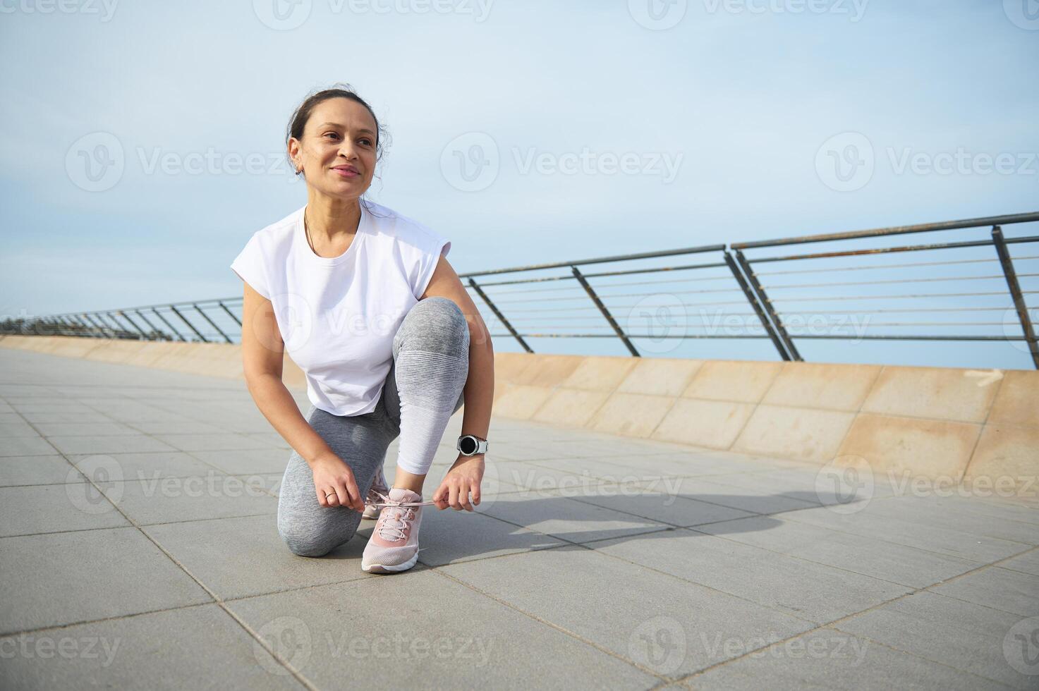 Happy woman runner tying laces on sports shoes, smiling looking away, getting ready for morning run on the promenade photo