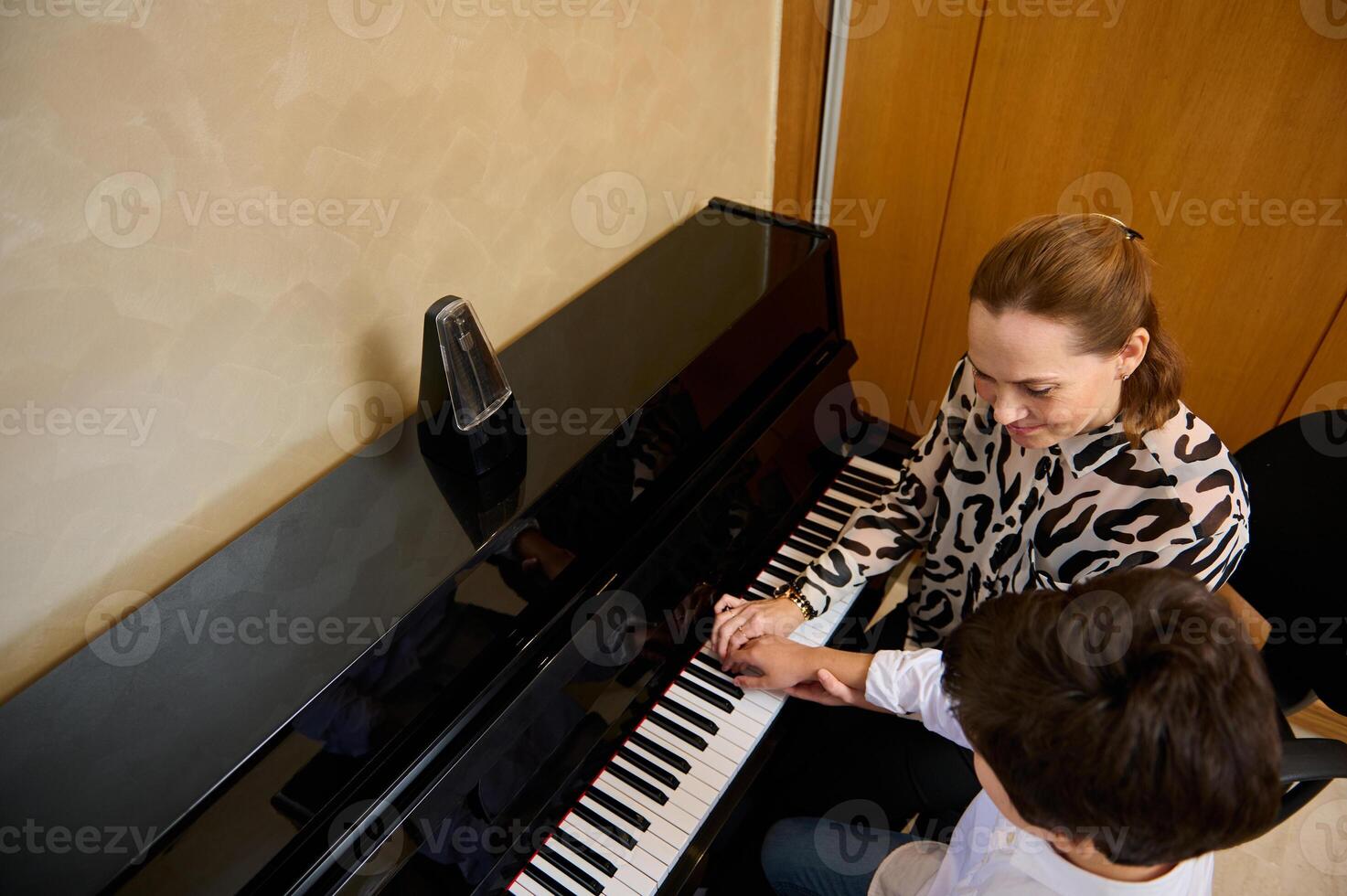 View from above of a Caucasian pleasant mature female music teacher, musician pianist teaching piano, explaining to a teenager student boy the correct position of fingers on the piano keys photo
