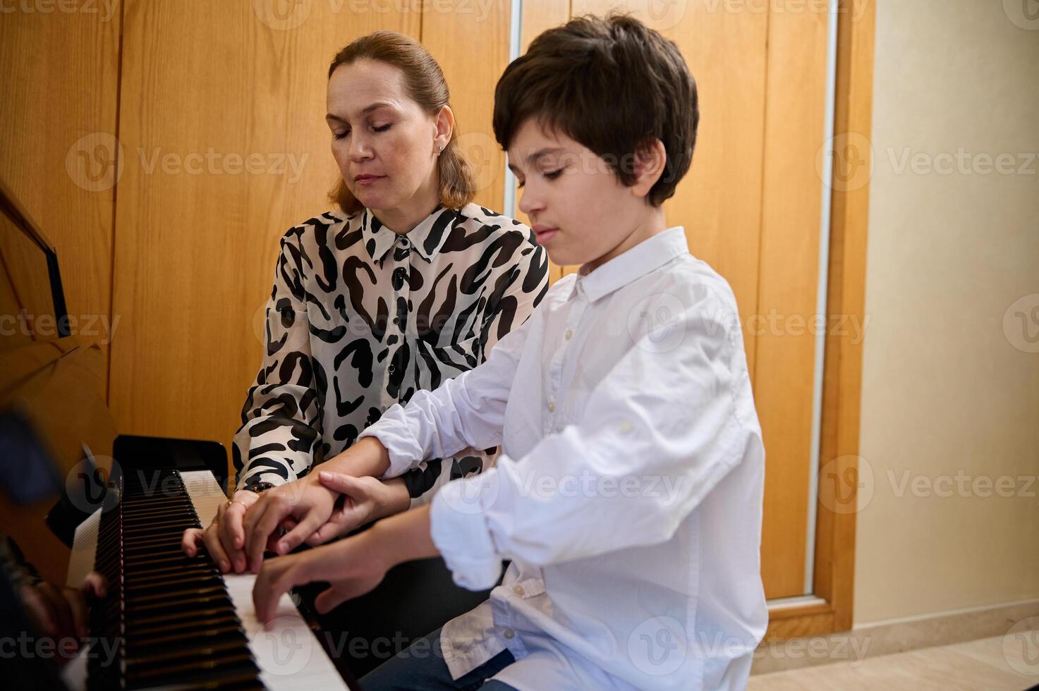 Handsome Hispanic boy in white casual shirt, learning playing piano, sitting at wooden piano forte and performing a musical composition under the guidance of is teacher. Musical education photo