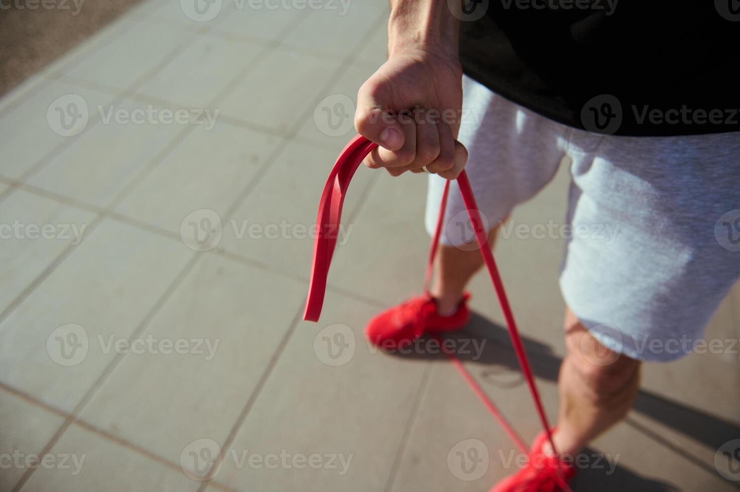 Top view of sportsman in red sneakers, gray sports shorts and black t-shirt doing warm up exercises with resistance band photo