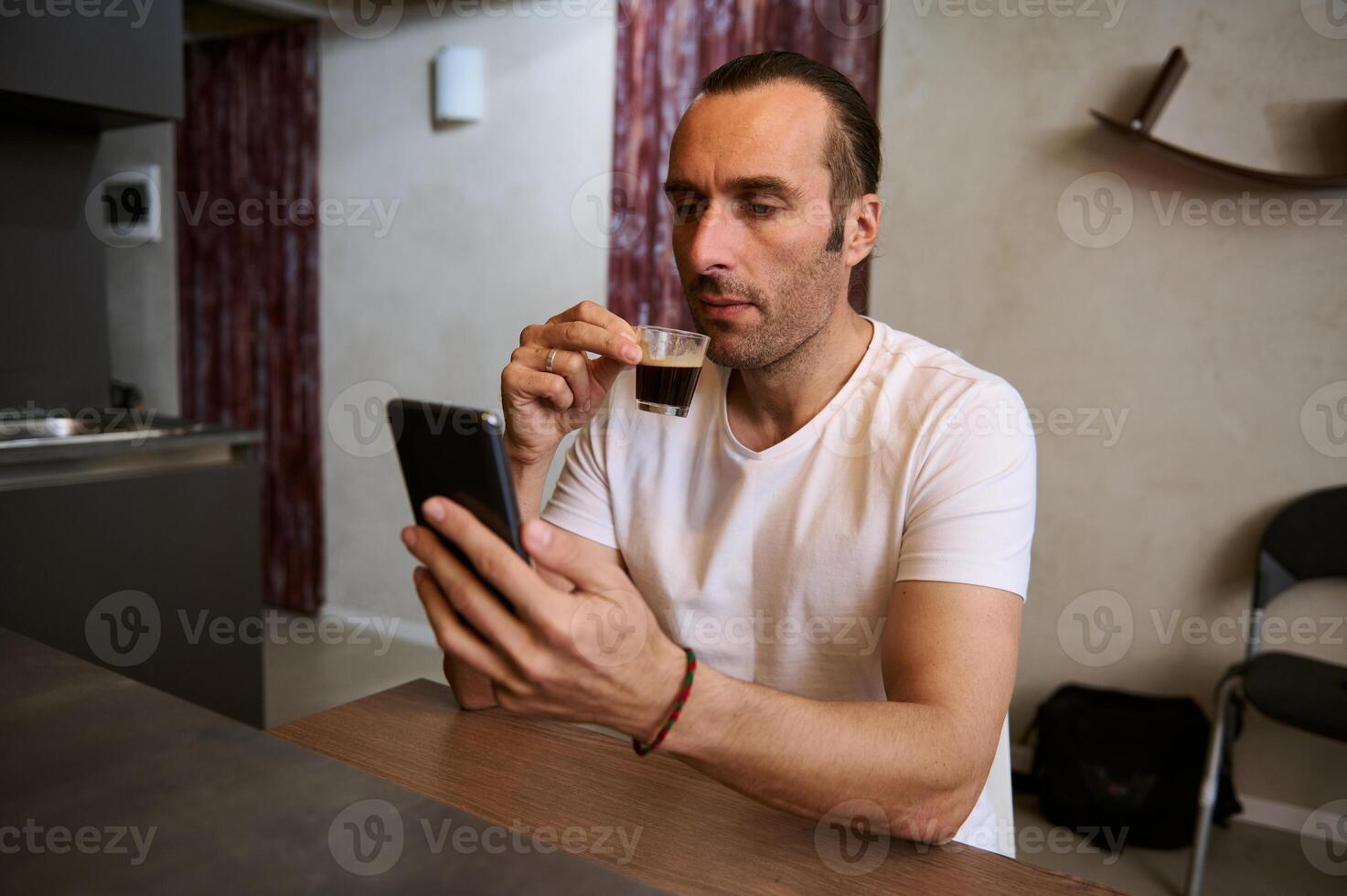 Authentic portrait of young Caucasian handsome man drinks coffee and reads news, sitting at table in the home interior photo