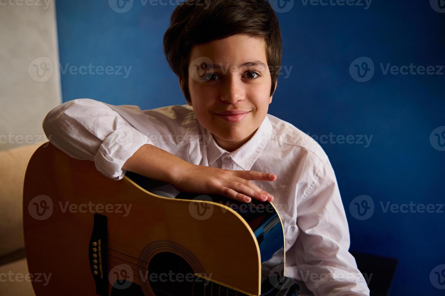 adolescente chico, un pequeño guitarrista músico participación acústico guitarra, sonriente mirando a cámara, sentado en contra un azul pared antecedentes en el música estudio foto