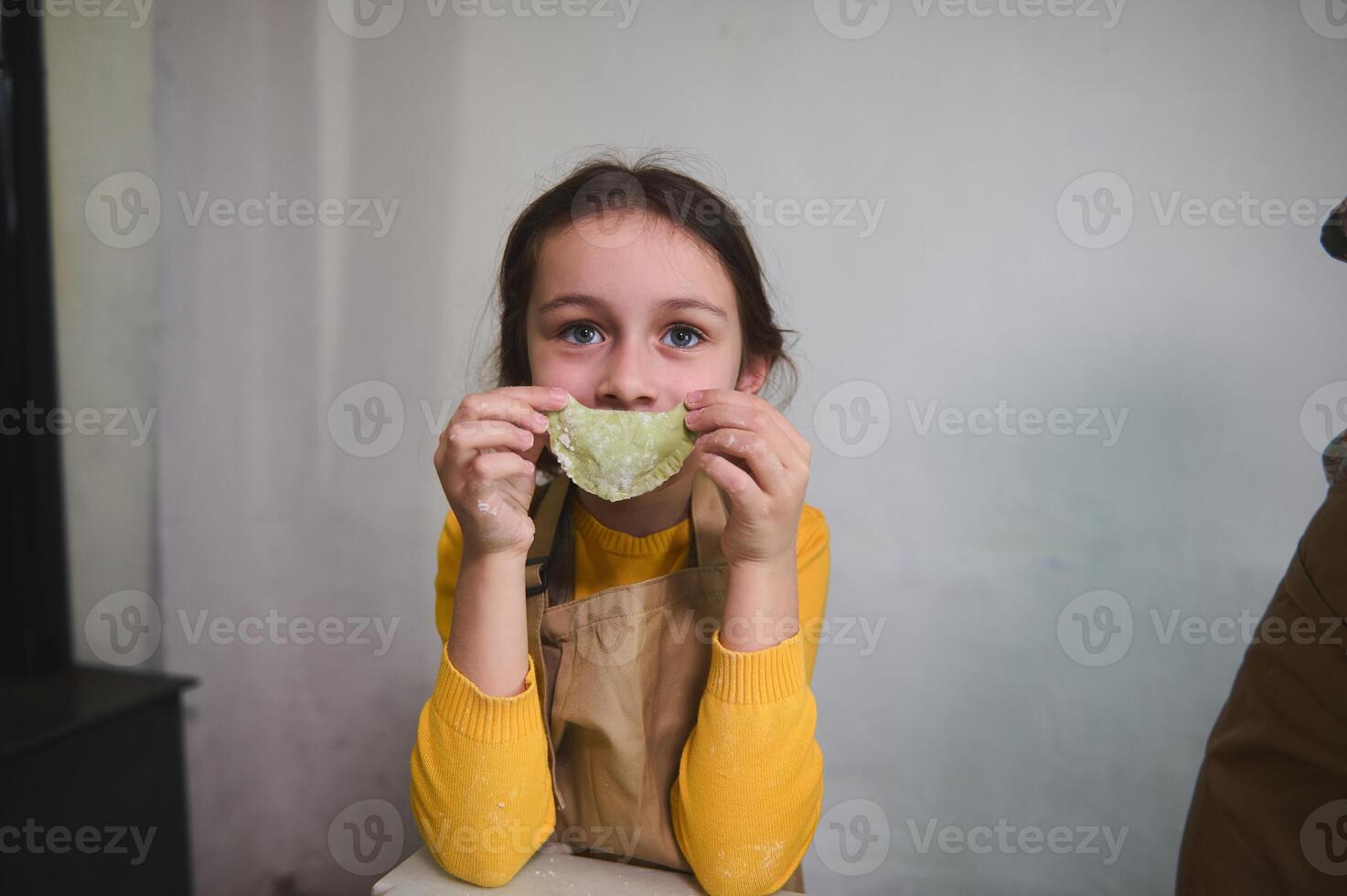 Portrait of a mischievous little girl in chef apron, holding a molded dumpling at the level of her mouth, making a beautiful smile looking at camera, standing against a white wall of a rustic kitchen photo