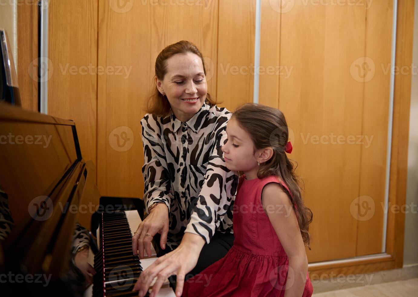 Cheerful mother and daughter pianists playing piano together, performing a melody for Christmas during music lesson together. Cute baby girl in elegant red dress, listens to her mom playing pianoforte photo