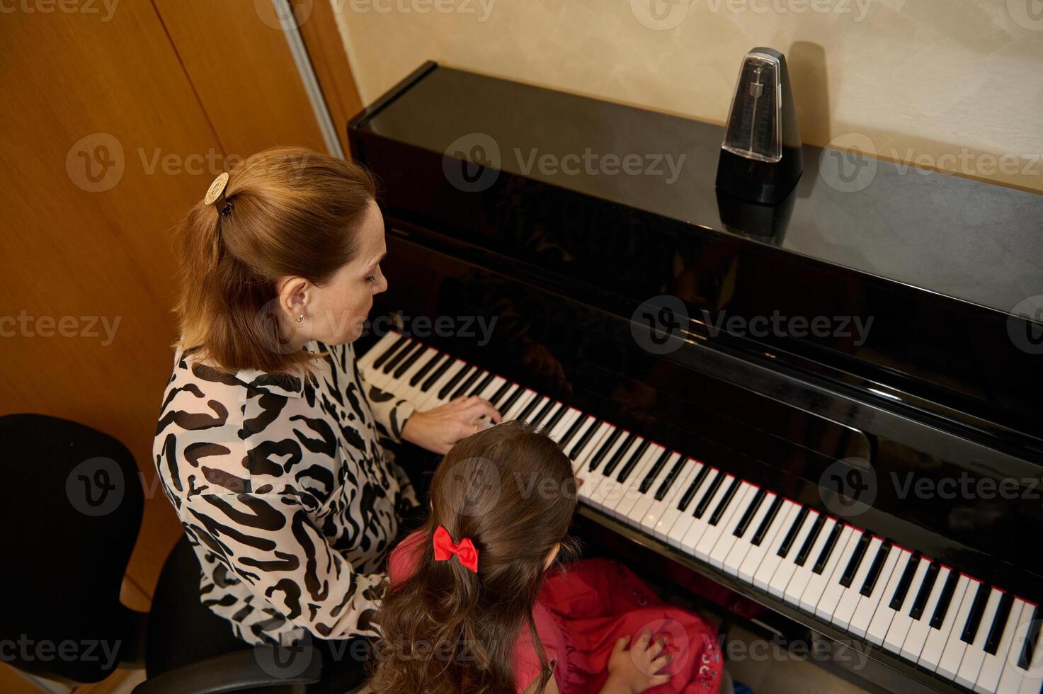 Rear view from above of a little child girl learning playing piano, touching piano keys under the guidance of her teacher during individual music lesson indoor. Art, culture and entertainment photo