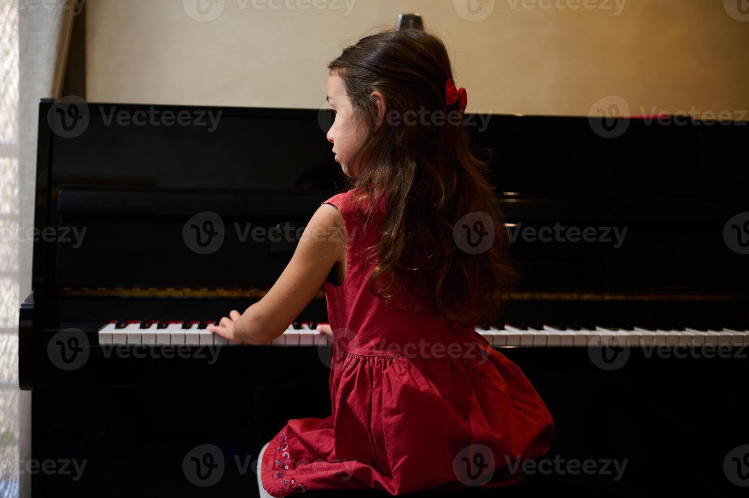 Authentic Caucasian little kid girl musician pianist with beautiful long hair, dressed in elegant red dress, putting fingers on piano keys, enjoying the performance of classical music. Copy ad space photo