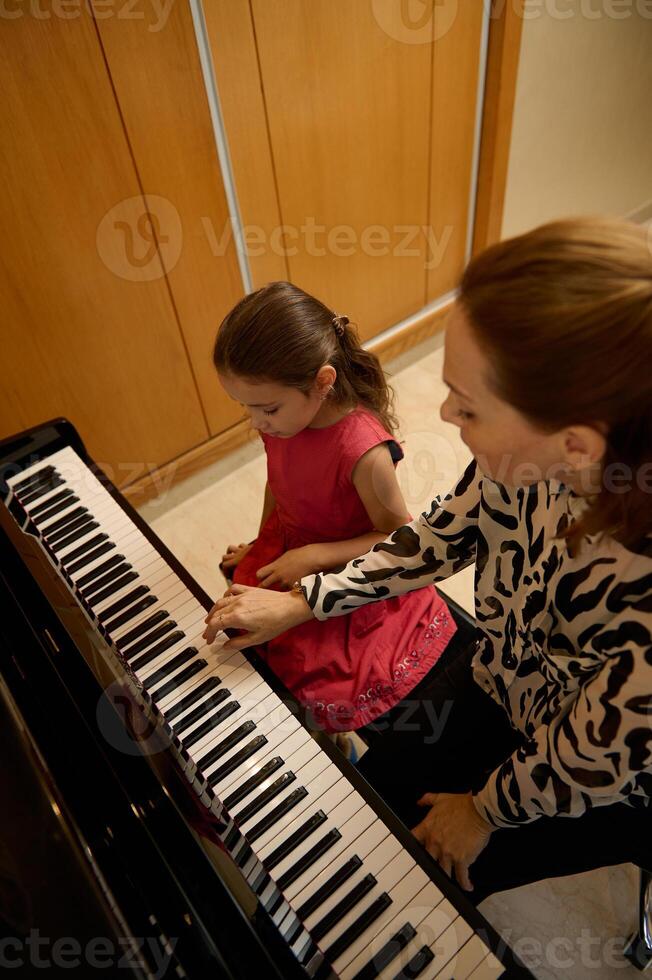 Overhead view of a female pianist musician holding the hands of a little child girl, showing the true position of finger on piano keys, explaining the piano lesson during individual music class photo