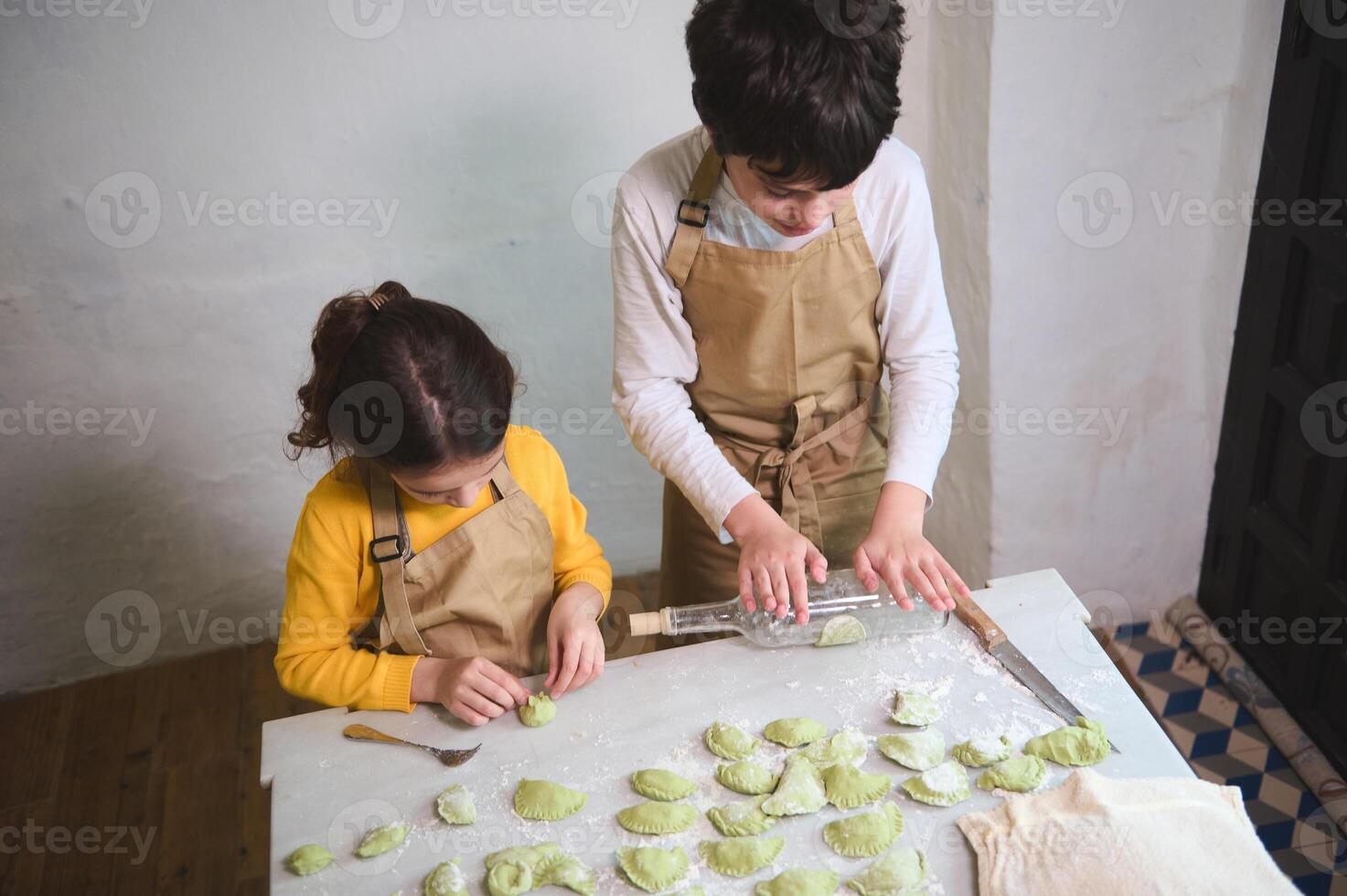 linda niños en el rural cocina, laminación fuera masa con un vino botella, esculpir empanadillas con machacado patatas relleno. Cocinando hecho en casa vegetariano albóndigas, italiano Ravioles o ucranio varenyky foto