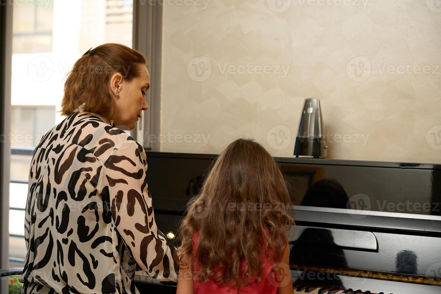 Rear view young adult woman, music teacher with the pupil, sitting at vintage wooden piano at home, giving a music lesson. Mother and cute daughter playing piano together. People. Hobbies and leisure photo