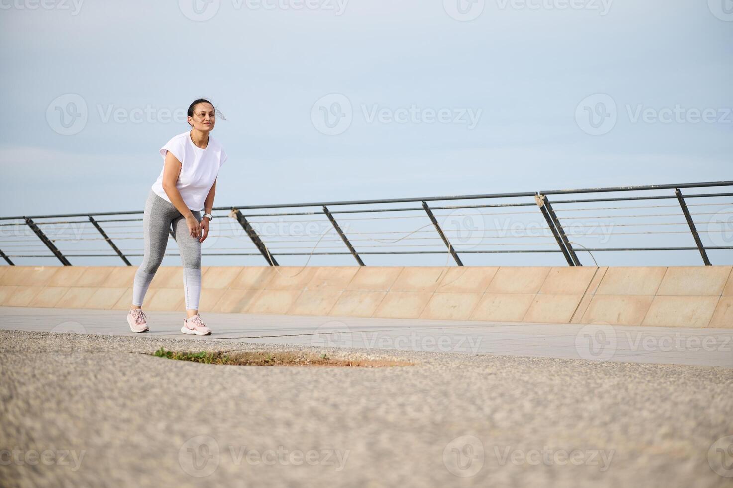 Full length shot of an exhausted young woman in sportswear running on the city bridge road photo