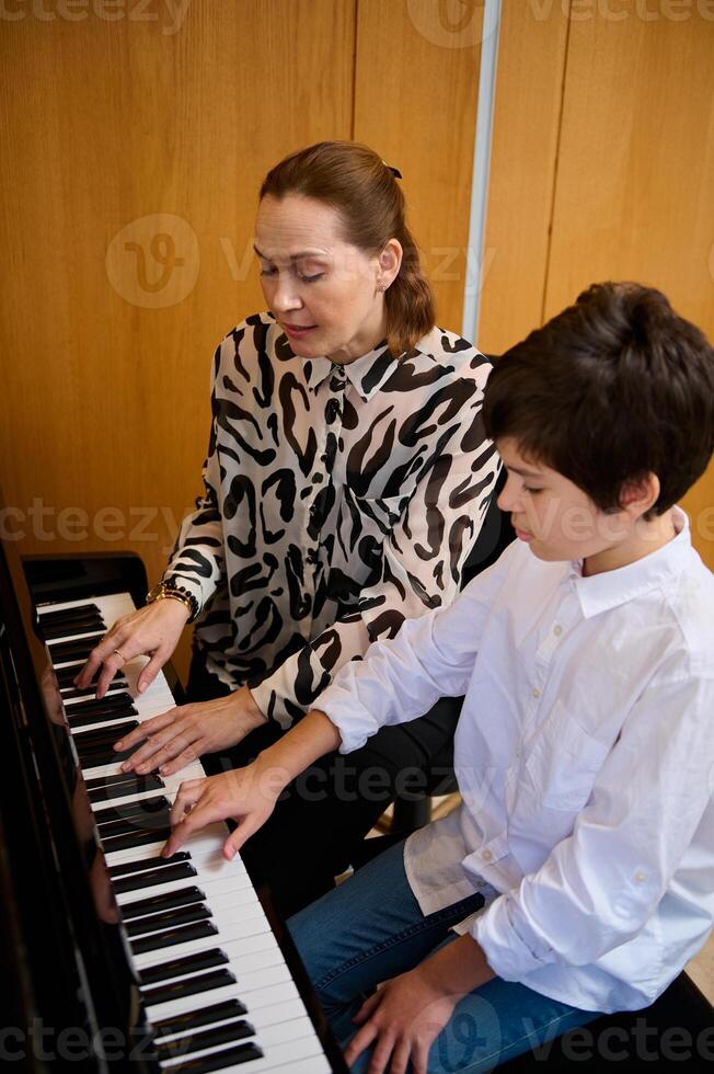 Vertical portrait of an authentic teenager boy having music lesson at home, sitting near his pianist teacher, playing piano forte photo