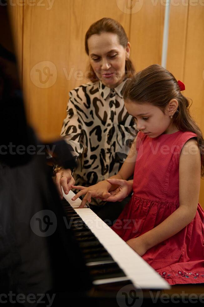 Vertical closeup portrait of a cute little girl in elegant red dress, sitting at wooden piano, with teacher learning to play piano indoors photo