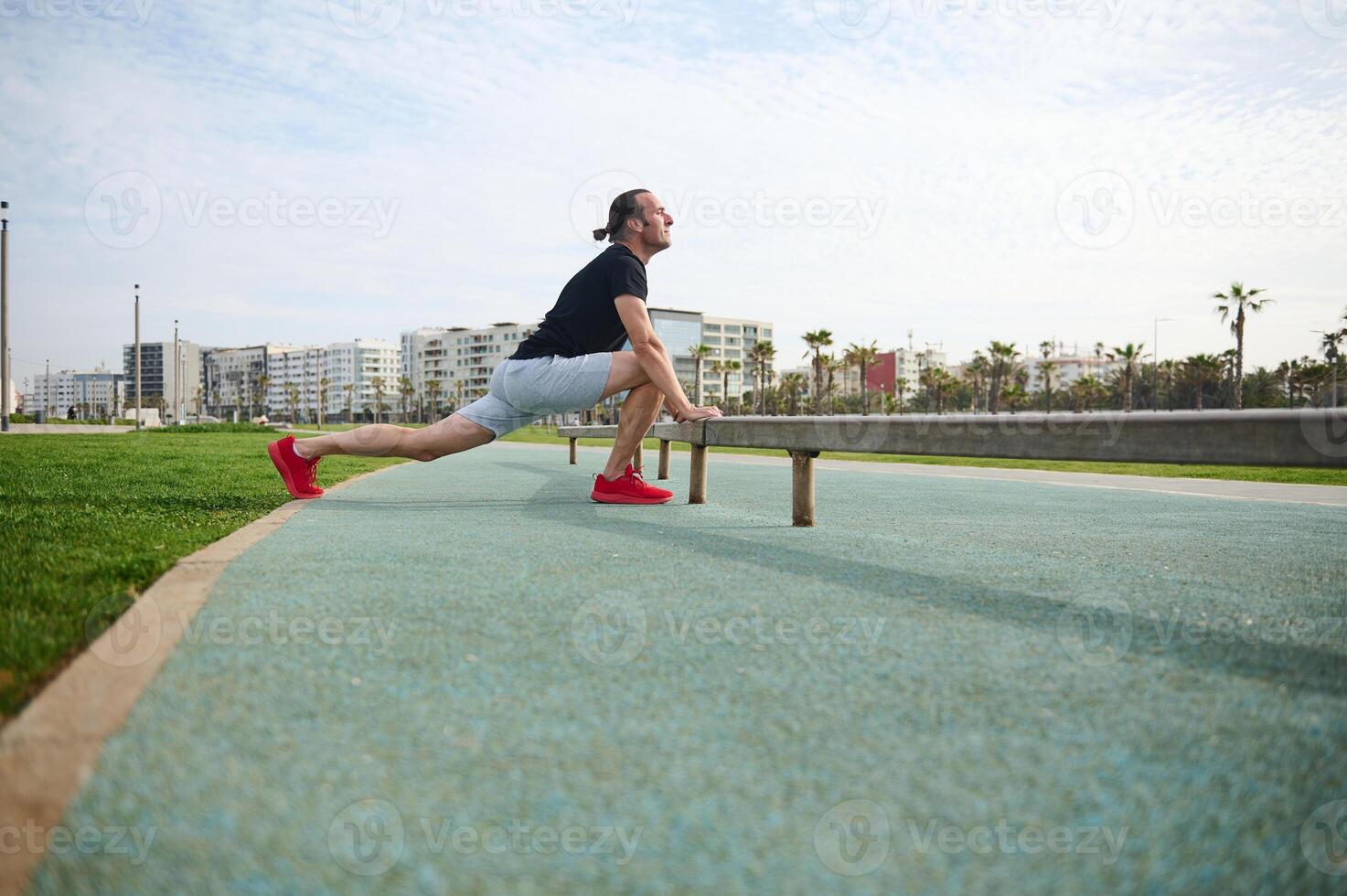 Determined sportsman in black t-shirt and gray sports shorts, warming up, doing lunges for stretching leg muscles. photo