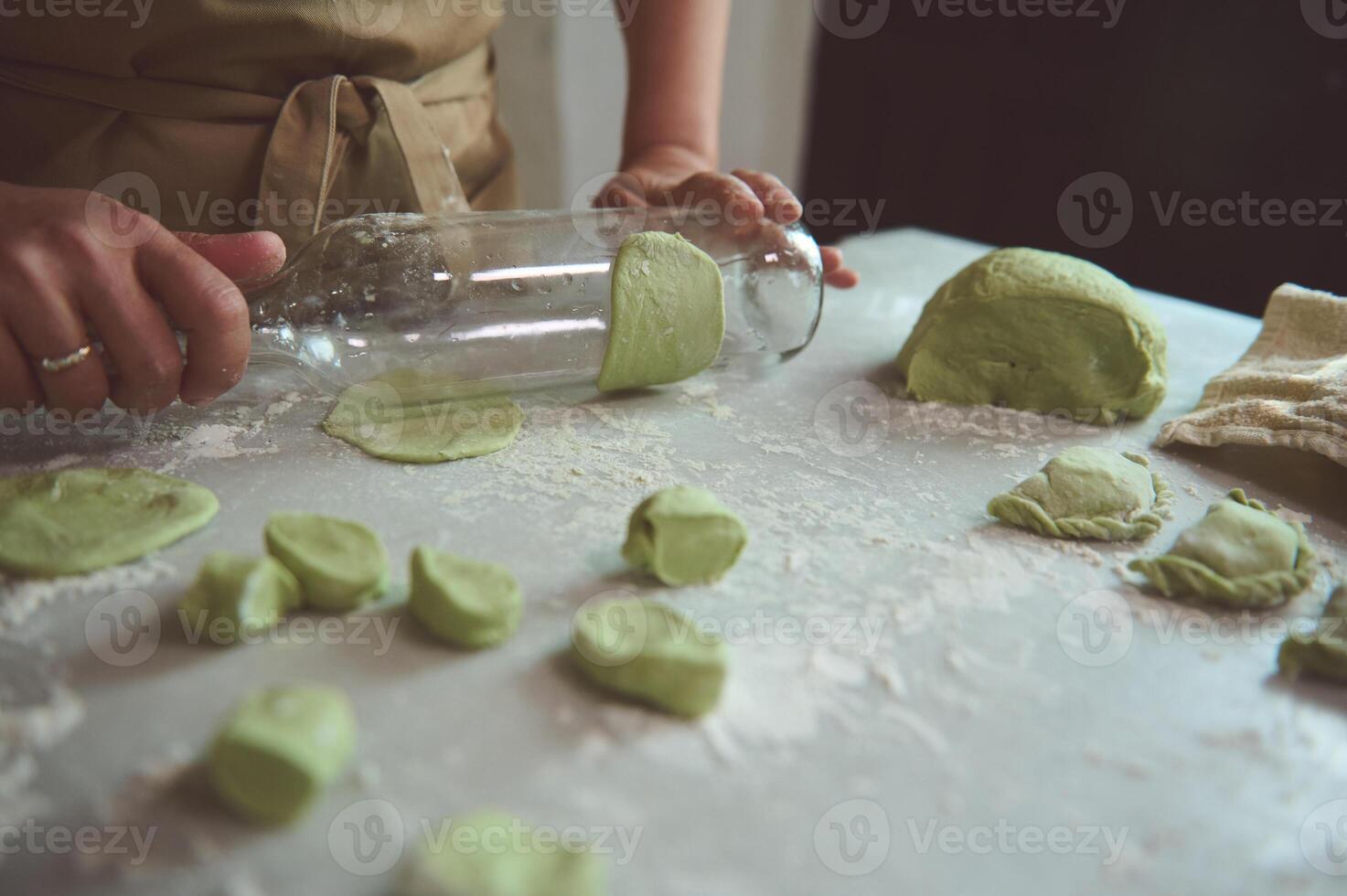 Close-up housewife, woman chef in beige apron, using an empty glass wine bottle as rolling pin, rolling out dough while cooking dumplings at rustic home kitchen. Female chef preparing green ravioli photo