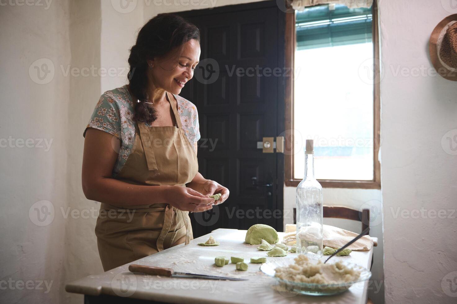 Authentic portrait of multi ethnic young charming woman, housewife in beige apron, standing at kitchen table with ingredients and spinach dough, modeling dumplings in rural kitchen rustic interior photo