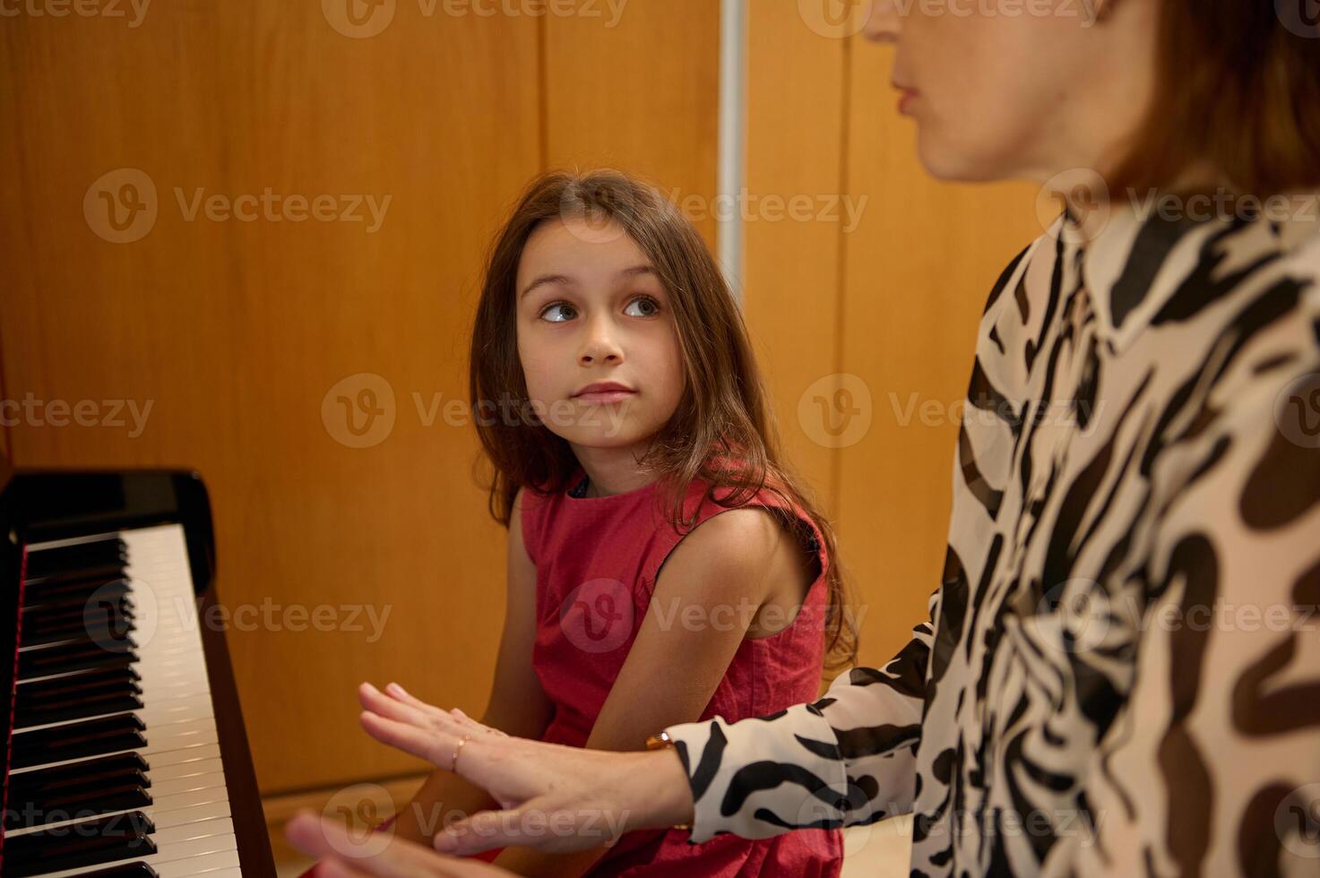 Authentic portrait of beautiful Caucasian elementary age schoolgirl in stylish red dress, smiling looking at her music teacher, sitting together at piano during individual music lesson indoors photo