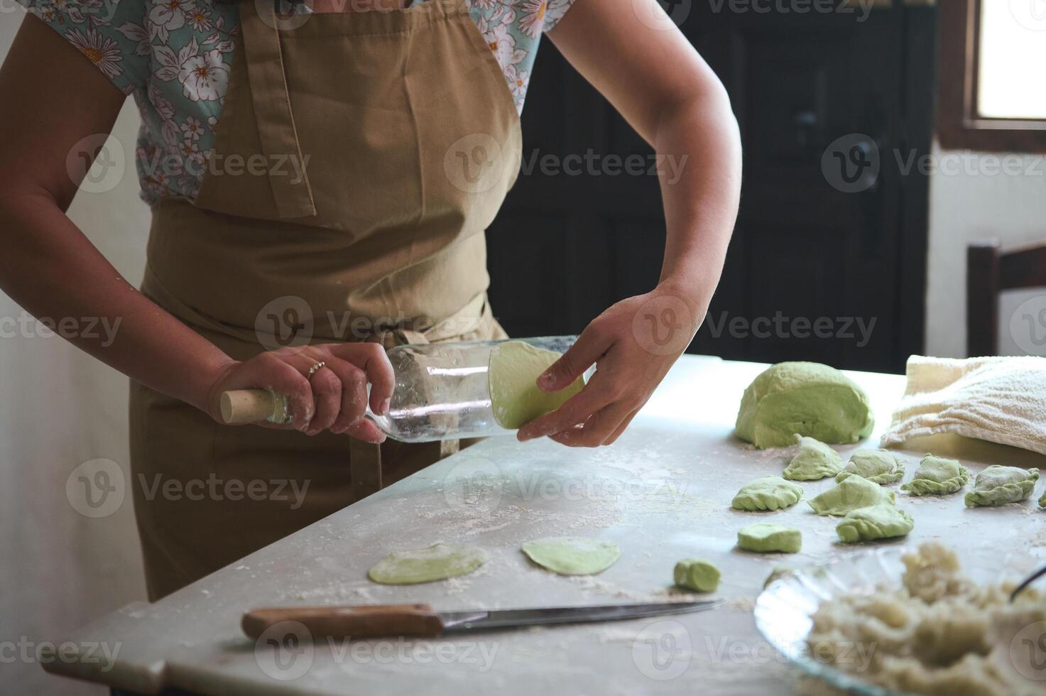 Close-up housewife using glass wine bottle as rolling pin, rolling out green spinach dough for making dumplings, standing at kitchen table sprinkled with flour in the rural house kitchen photo