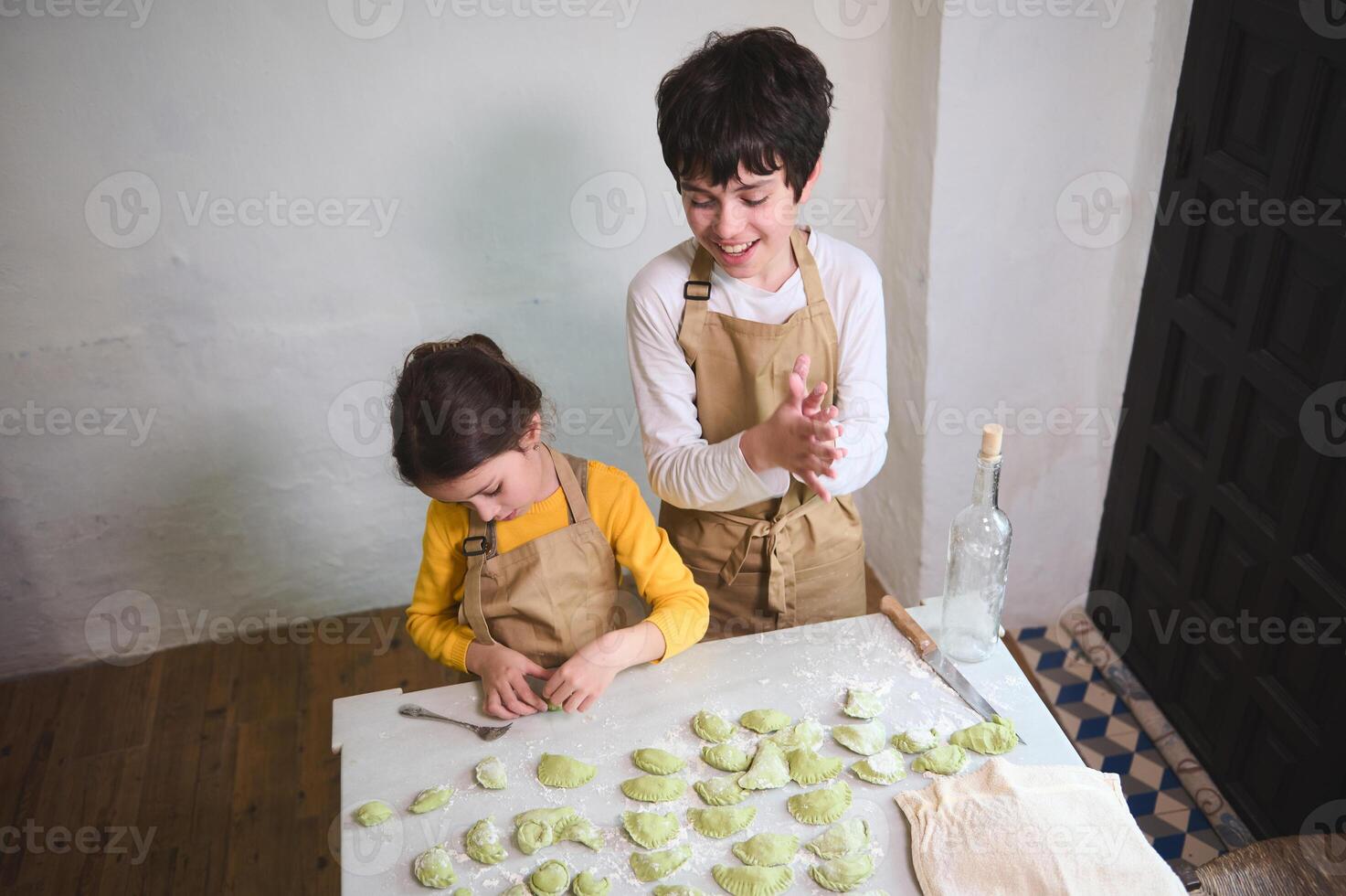 Cooking class for kids. Two kids, boy and girl preparing family dinner, standing at floured kitchen table and modeling dumplings or Ukrainian varennyky in the rural house kitchen interior. Top view photo