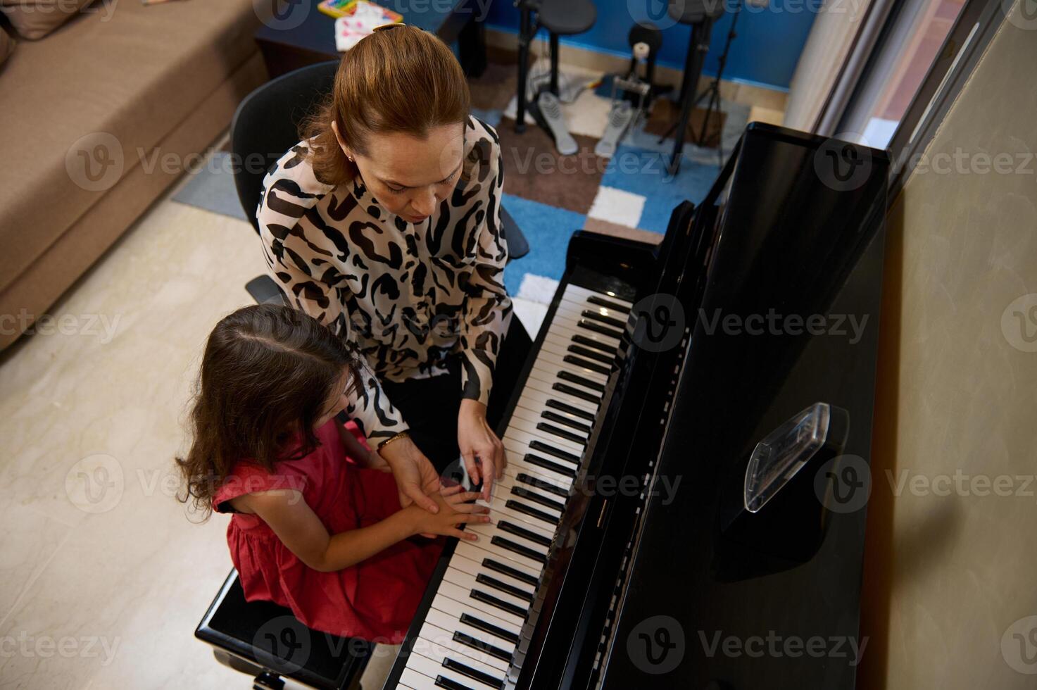 caucásico agradable maduro hembra música maestro, pianista enseñando piano, explicando a un pequeño niño niña estudiante el correcto posición de dedos en el piano llaves. ver desde encima foto