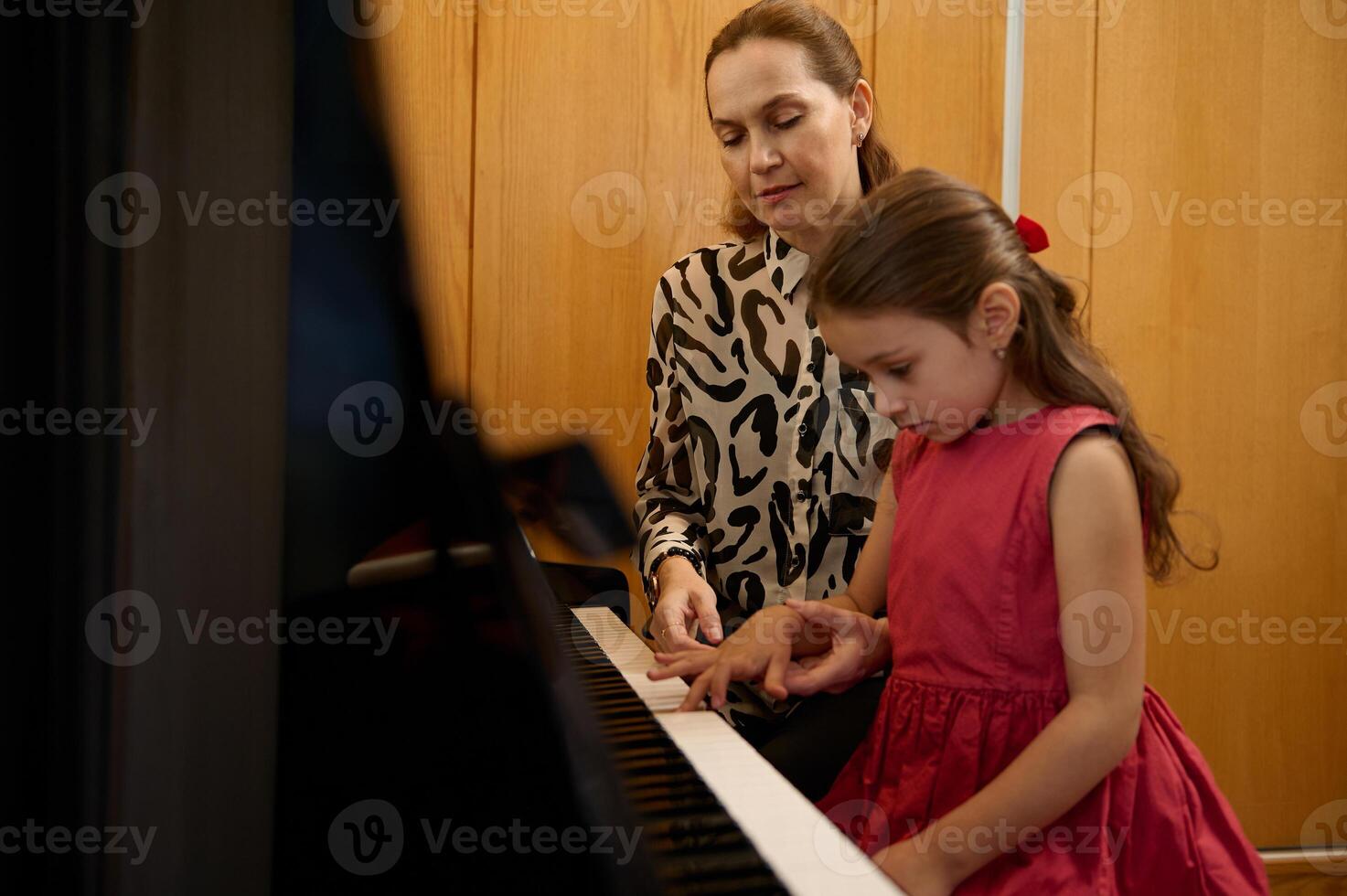 Adorable little girl in red dress, taking piano lesson, passionately playing the keys under her teacher's guidance, feeling the rhythm of music. Musical education and talent development in progress photo