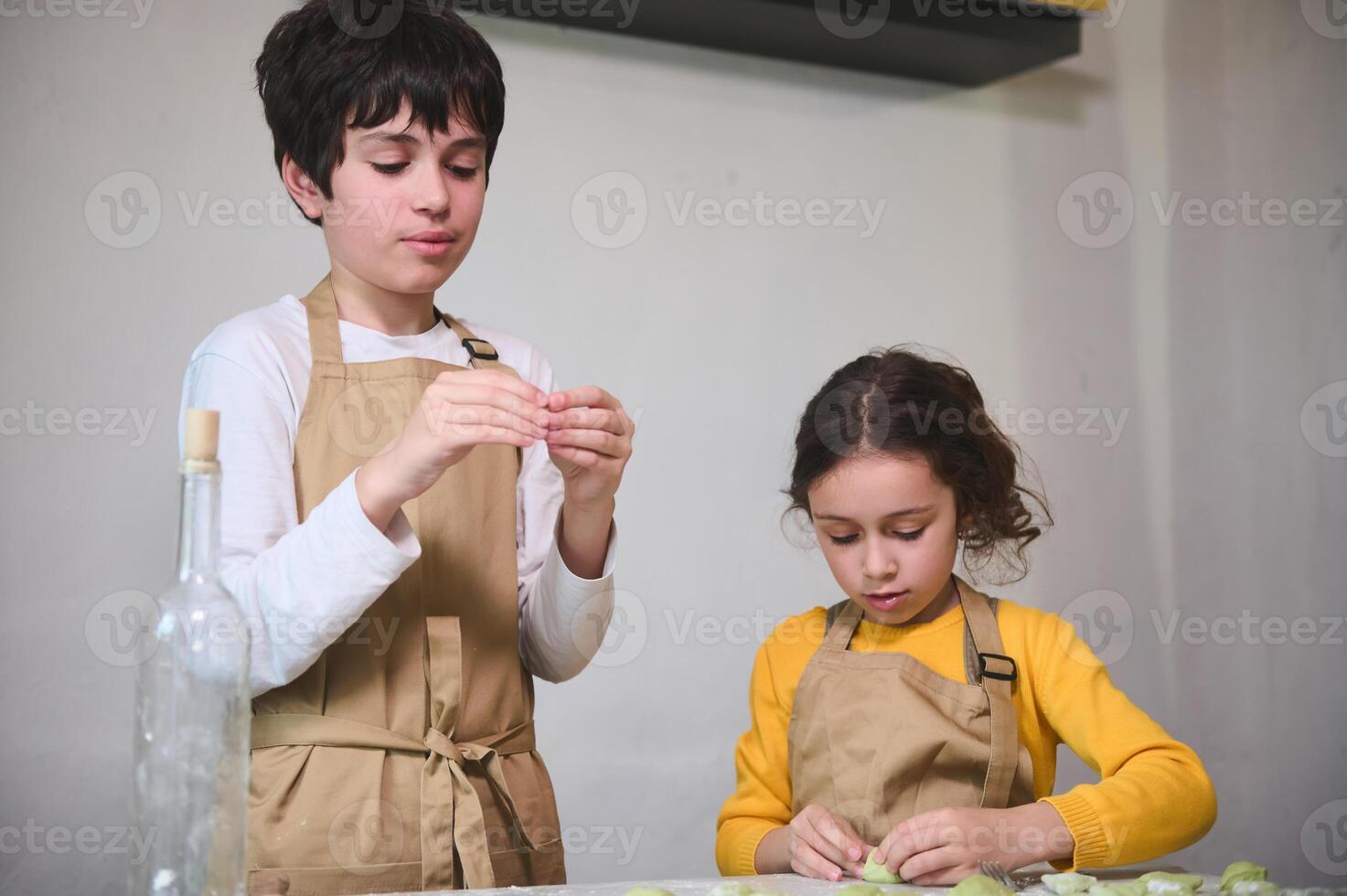 Cute children in beige chef apron, preparing dumplings during a cooking class indoors, standing against a white wall background photo