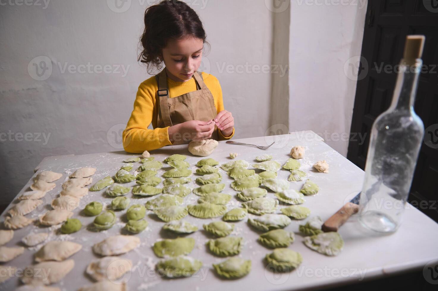 encantador pequeño niño niña Relleno masa con machacado papas, modelado albóndigas, en pie a cocina mesa con amasado masa y ingredientes. el proceso de Cocinando Ravioles, varenniki, vegano pelmeni foto