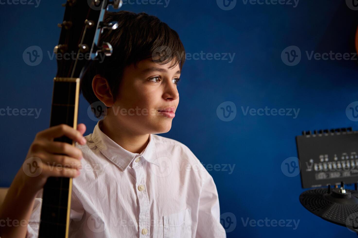 adolescente chico músico guitarrista en blanco camisa, soñando mirando aparte, posando con acústico guitarra azul pared antecedentes foto