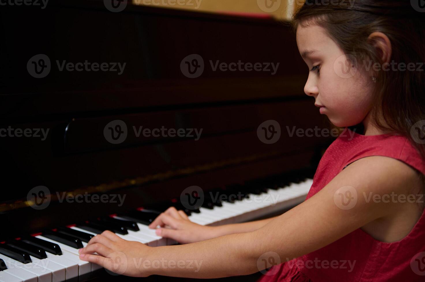 Authentic portrait of a Caucasian little kid girl in elegant red dress, sitting at piano forte, performing classical melody, feeling rhythm of music, touching ivory and ebony keys of the grand piano photo