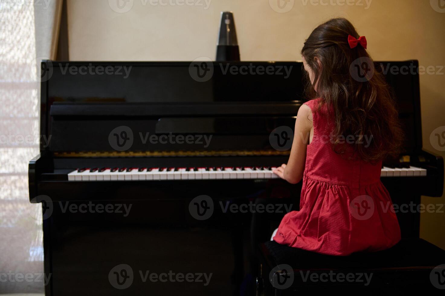 View from the back of little child girl in stylish red dress, playing grand piano at home. Talented kid, musician pianist composing a melody, putting fingers on black and white piano keys. Copy space photo