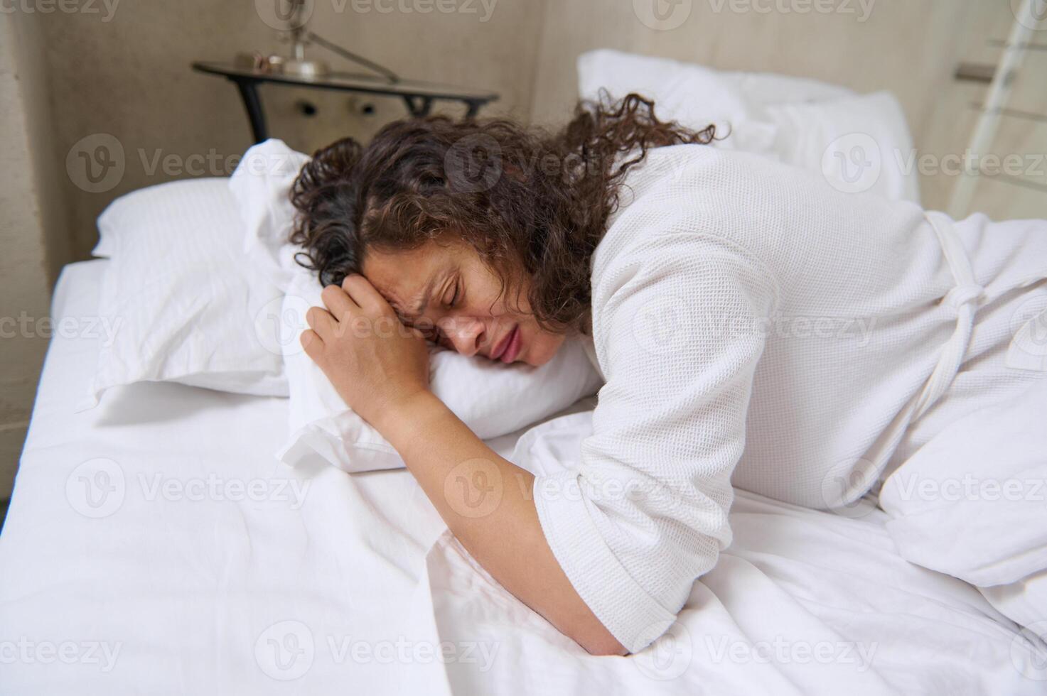 Young brunette crying and feeling depression, lying on white bed sheets in her bed in the home bedroom photo