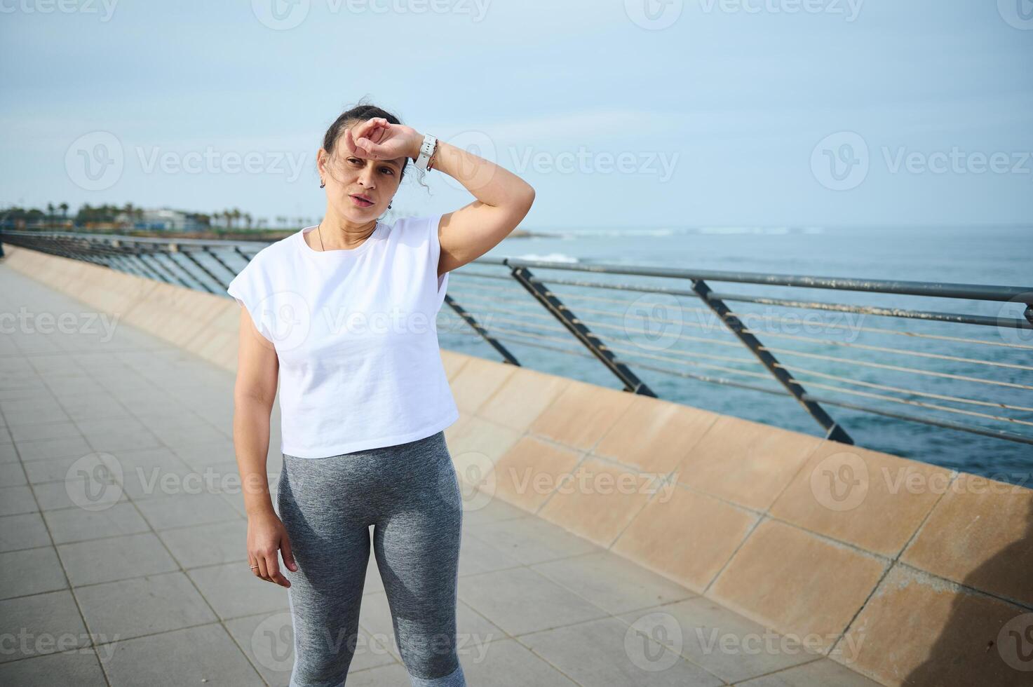 Exhausted young athletic woman runner wiping her forehead, standing on the bridge, relaxing after morning jog photo