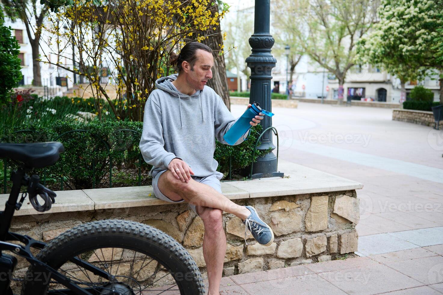 Athletic young man cyclist relaxing when riding electric bike, holding a bottle of water while sitting on the city bench photo