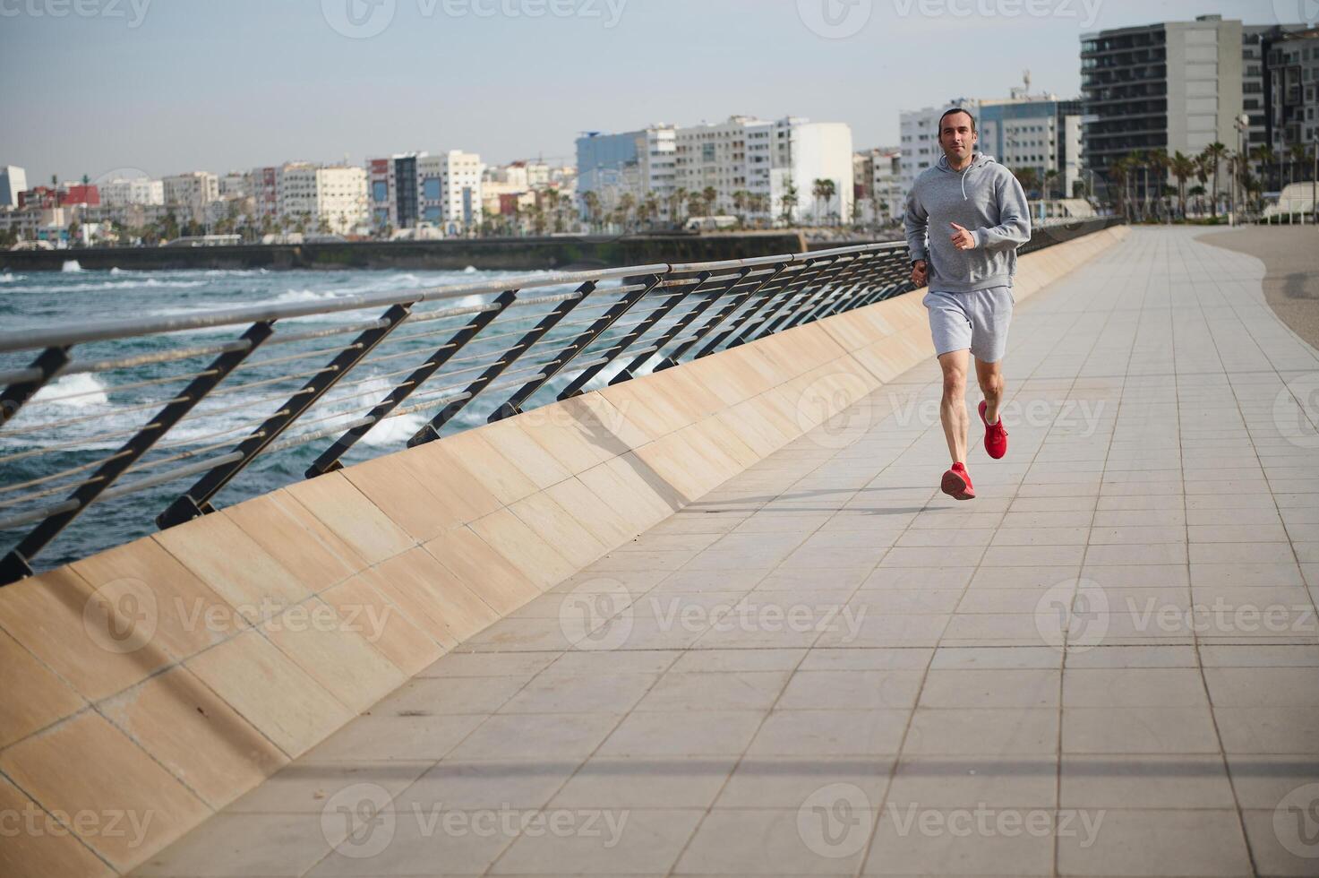 Full length portrait handsome young athletic man jogging, performing sprint run on running track in the coastline city photo