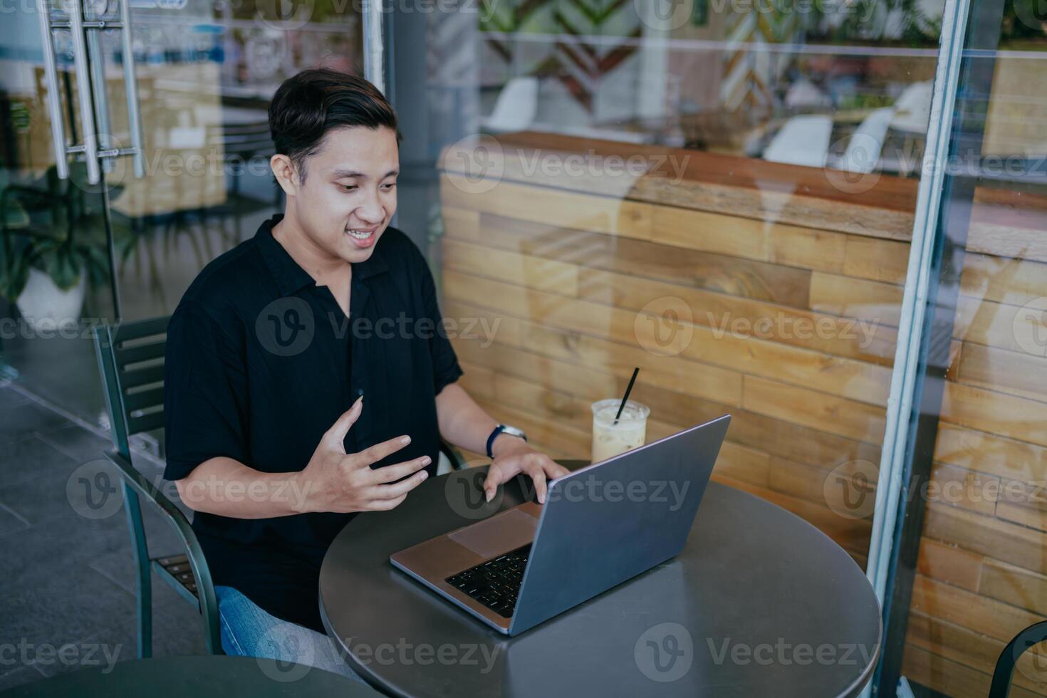 Handsome young man sitting in online meeting in outdoor cafe, talking to laptop camera, explaining something, drinking coffee. photo