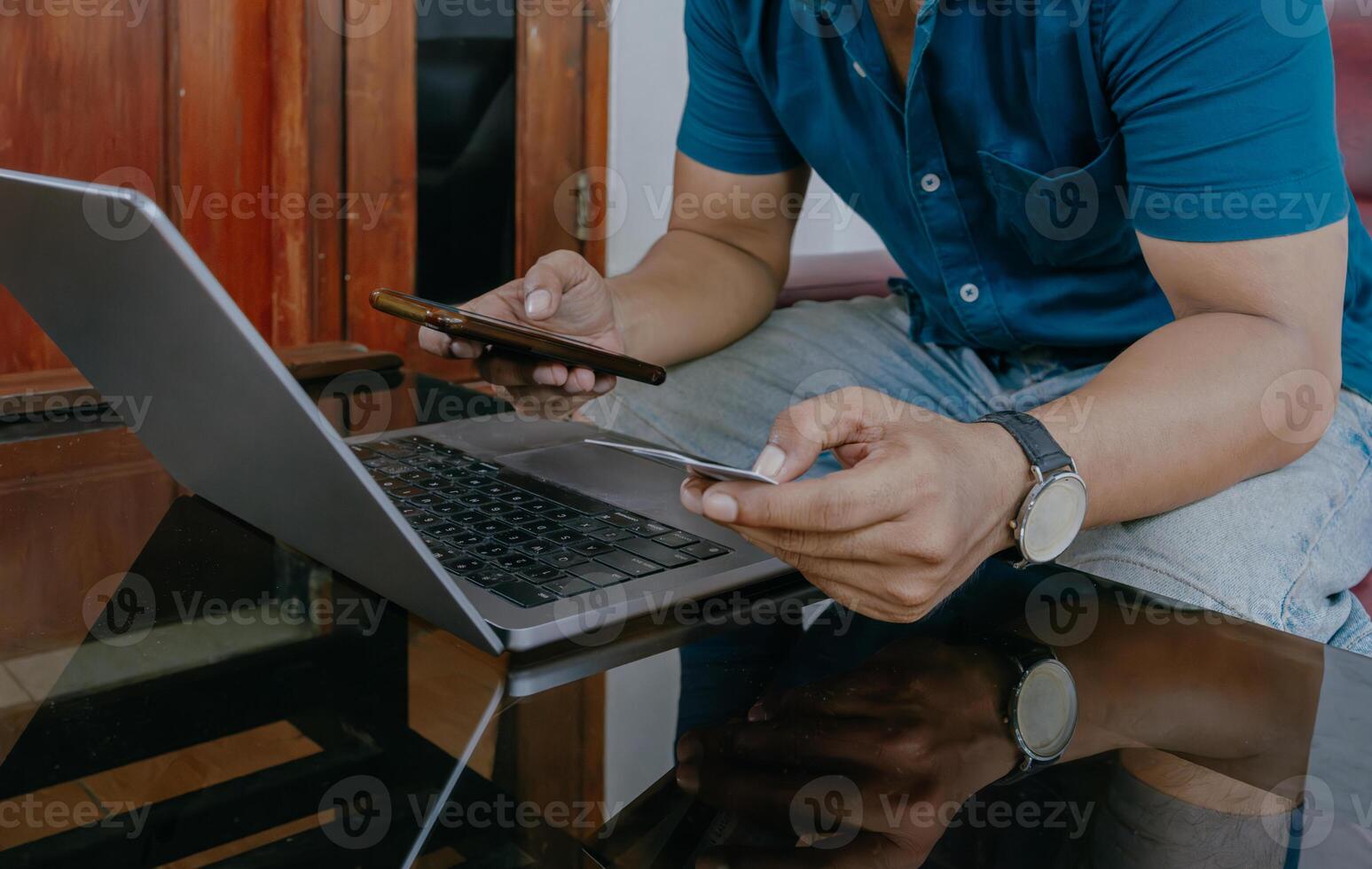 Close-up of man's hand holding cellphone and credit card. Man working using laptop at desk in home office. photo