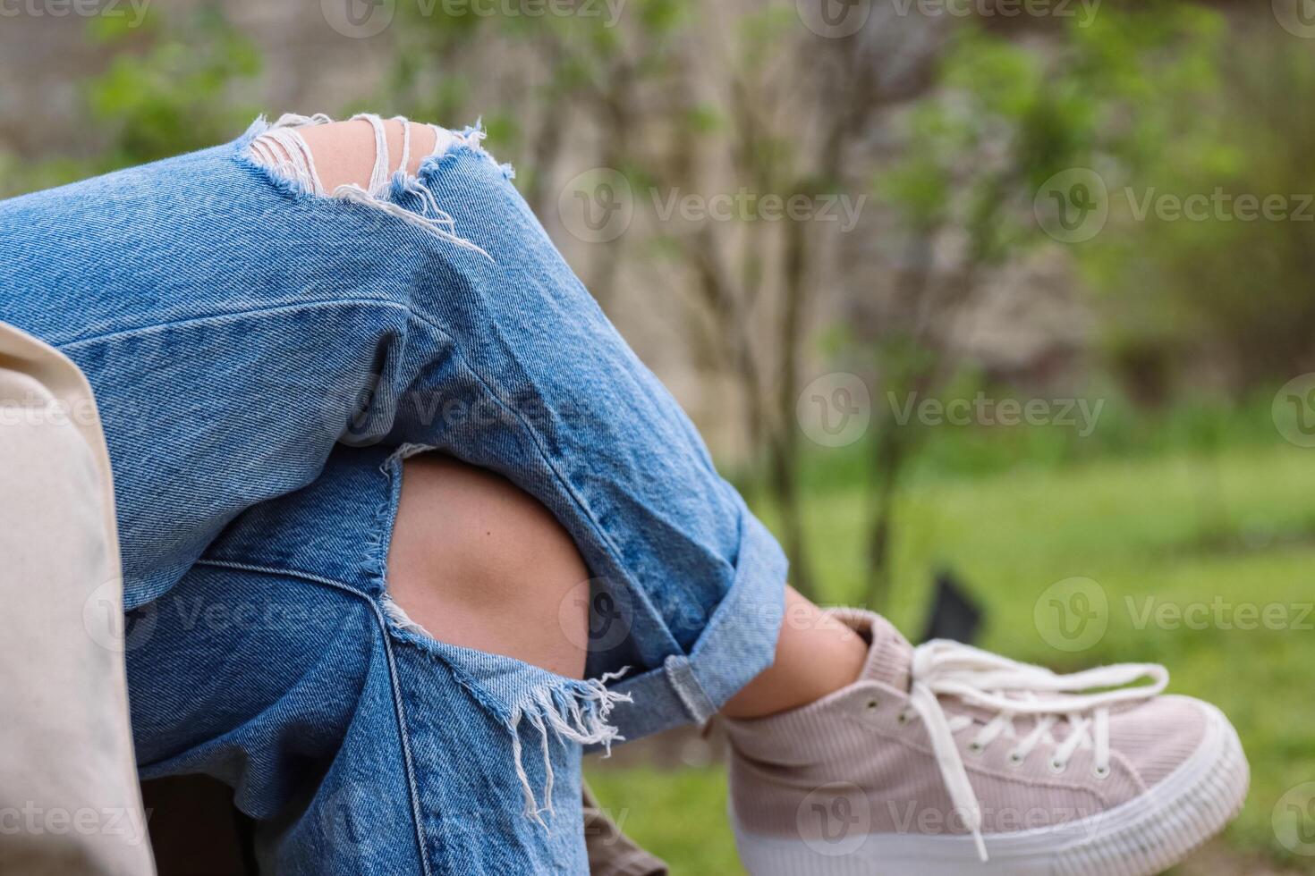 Young girl wearing ripped blue jeans with big hole on the knee is sitting on the bank crossed her legs. Woman in destroyed distressed denim. Close up of Textile texture. Selective focus, blurred photo