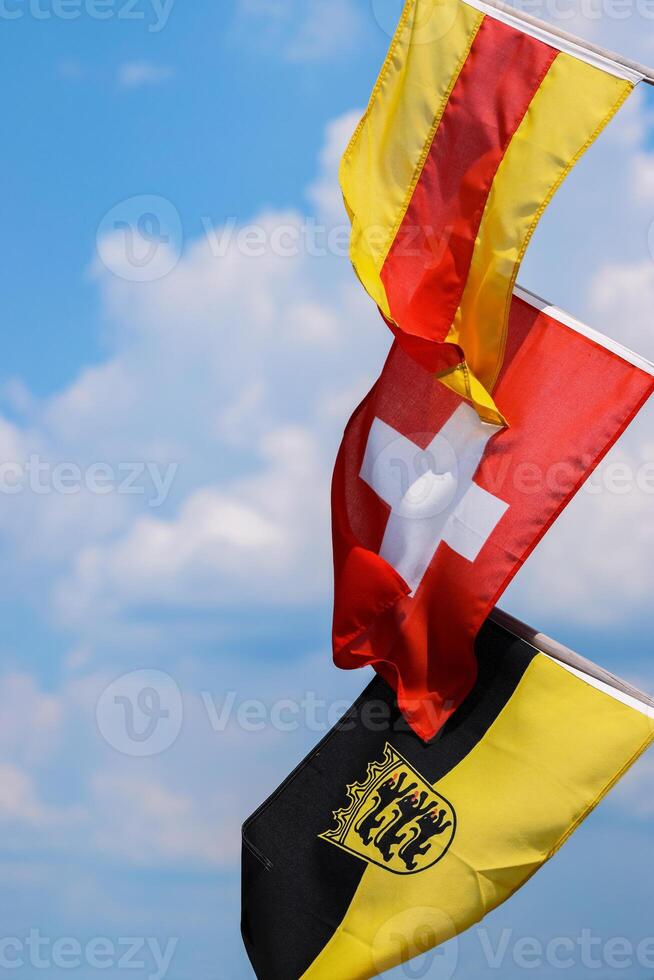 Three flags on the flagpoles waving in the wind. Flags of Baden, Switzerland, Flag Baden-Wuerttemberg with an emblem. Swiss flag in the middle. Isolated on the blue sky and white clouds. Copy space. photo