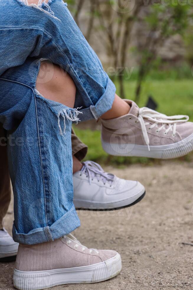 Two young girls wearing white sport shoes, ripped blue jeans with big hole on the knee sitting on the bank and crossed there legs. Woman in destroyed distressed denim. Selective focus, blurred photo