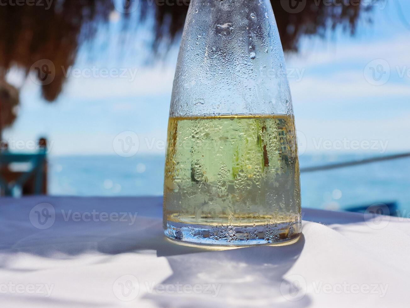 Fresh fruit water in a glass pitcher on the table in the restaurant at the sea shore. Ice cold lemonade at the beach. Detail of the bottle. Drops of water. Blue sky and sea on the background. Close-up photo